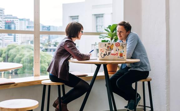 Two people sit at a high table, facing each other with a laptop between them.