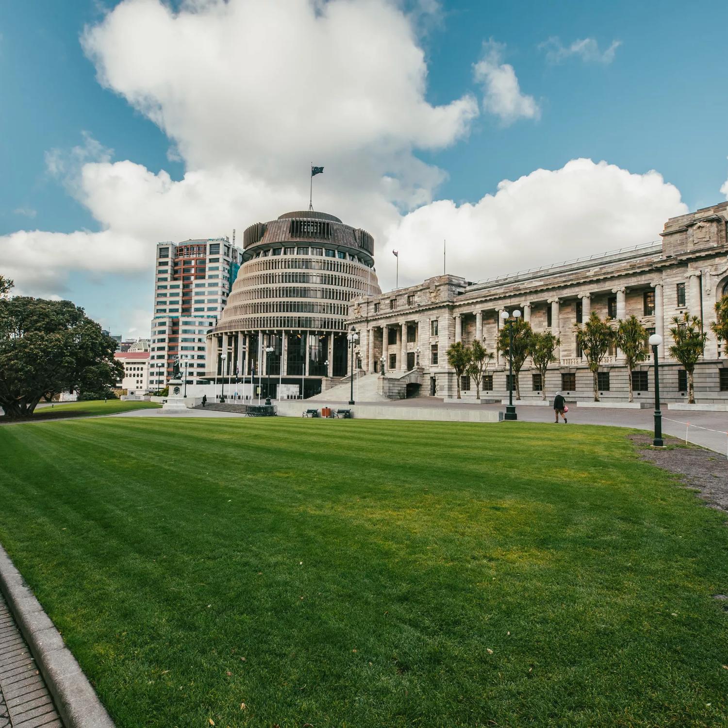 Looking towards the Beehive and Parliament buildings across a grass park, tress are surrounding and people walk by.