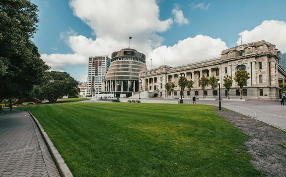 Looking towards the Beehive and Parliament buildings across a grass park, tress are surrounding and people walk by.