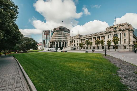 Looking towards the Beehive and Parliament buildings across a grass park, tress are surrounding and people walk by.