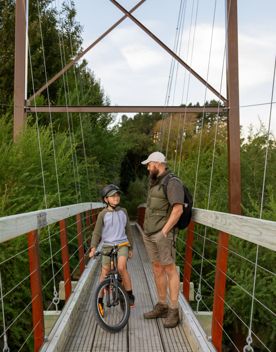 A father and child stand next to each other on a red swing bridge on the Tauwharenikau Trail in the Wairarapa region