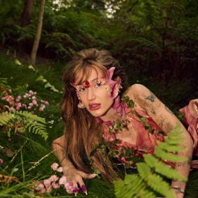 Australian pop singer Peach PRC wears a pink floral costume while lying on the forest floor.