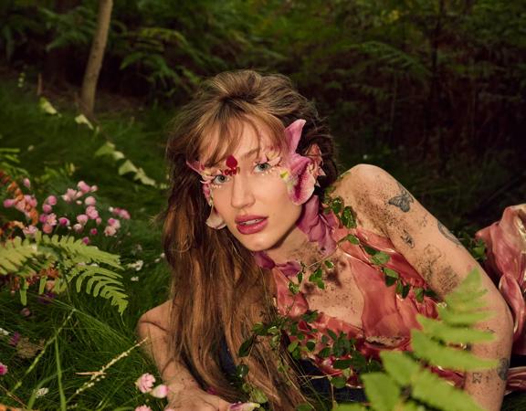 Australian pop singer Peach PRC wears a pink floral costume while lying on the forest floor.