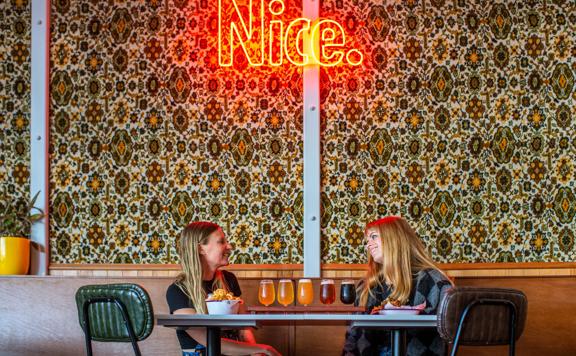 2 poeple share a beer tasting tray at Parrotdog Bar, with an Led sign above them that says Nice.