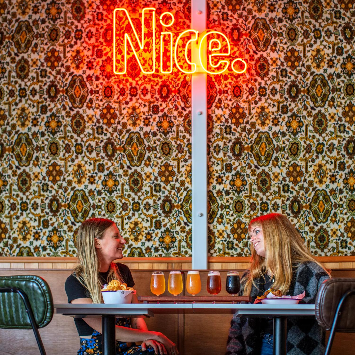 2 poeple share a beer tasting tray at Parrotdog Bar, with an Led sign above them that says Nice.