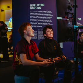 Visitors using game controllers and interacting with digital displays in a darkened exhibition space.
