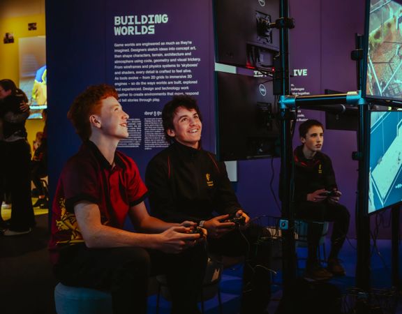 Visitors using game controllers and interacting with digital displays in a darkened exhibition space.