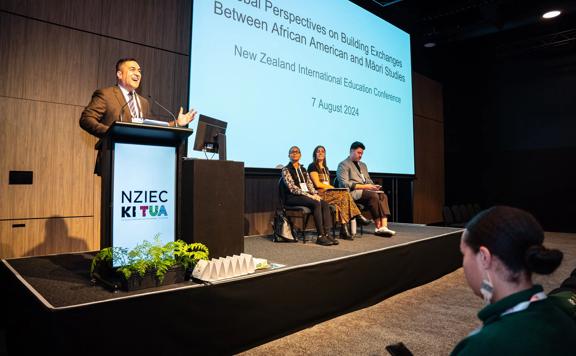 A person in a brown suit speaks at a podium at the New Zealand International Education Conference 2024, held at Tākina.