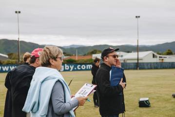 Behinds the scenes on the set of 'Extreme Cake Sports'. The producer, Bevin Linkhorn, wears a black jacket, black cap and sunglasses is holding a blue clipboard with is right arm and has his left hand near his mouth to amplify his voice.