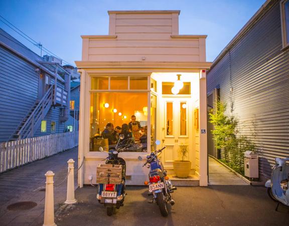 The exterior of Rita in Te Aro valley at sunset with their yellow lighting shining through the glass.