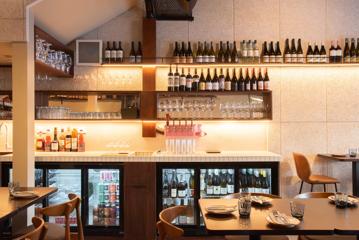 Rosella Wine Bar interior with bottles on shelfs along a grey wall, a white counter with drink fridges and tables set for dining in the foreground.