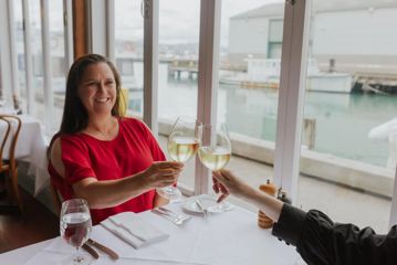 A person reaches out with their white wine to cheers with a friend at a window-side table.