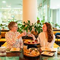 Two people clink wine glasses at high tea at t-Lounge by Dilmah. A cake stand filled with food sits on the table in front of them.