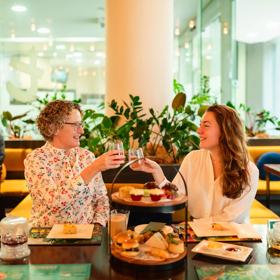 Two people clink wine glasses at high tea at t-Lounge by Dilmah. A cake stand filled with food sits on the table in front of them.