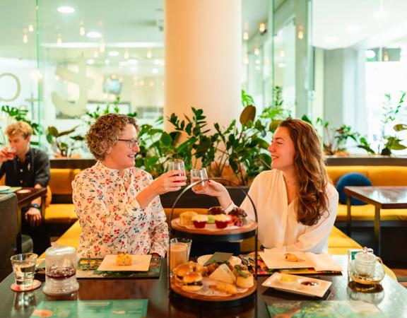 Two people clink wine glasses at high tea at t-Lounge by Dilmah. A cake stand filled with food sits on the table in front of them.