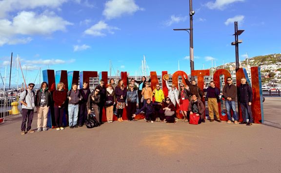 A large group of people stands in front of the red “Wellington” sign along the waterfront, with boats, buildings, and a blue sky in the background.