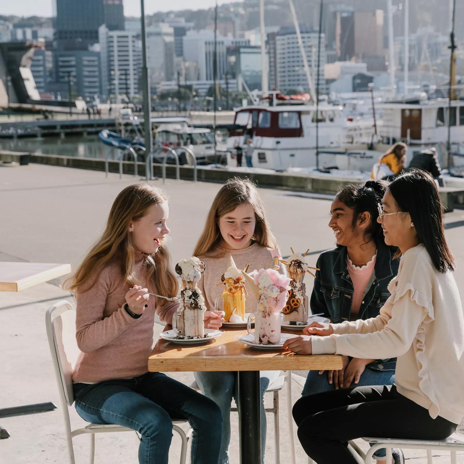 4 young people sit outside The Enormous Crocodile & Shake Bar, each enjoying a large milkshake with wacky toppings.