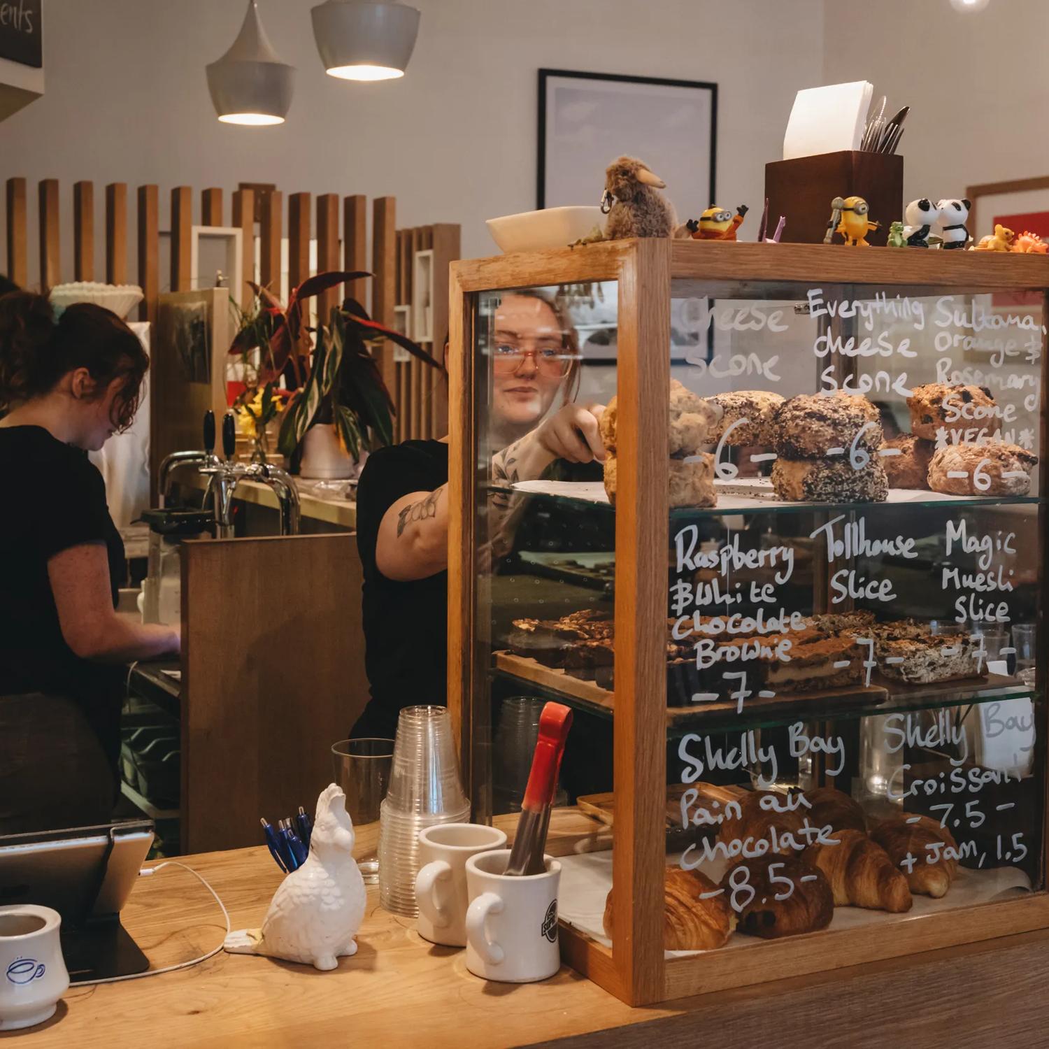 The front counter at Neo Café & Eatery with three workers behind the counter, one of which is grabbing a scone from the display window. 