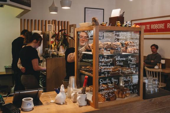 The front counter at Neo Café & Eatery with three workers behind the counter, one of which is grabbing a scone from the display window. 