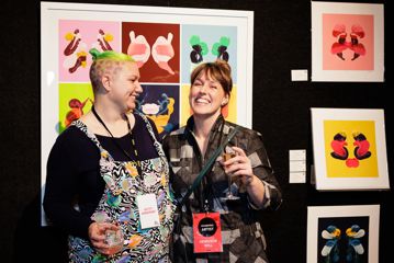 Two people smiling and holding drinks in front of brightly coloured artworks on a wall.