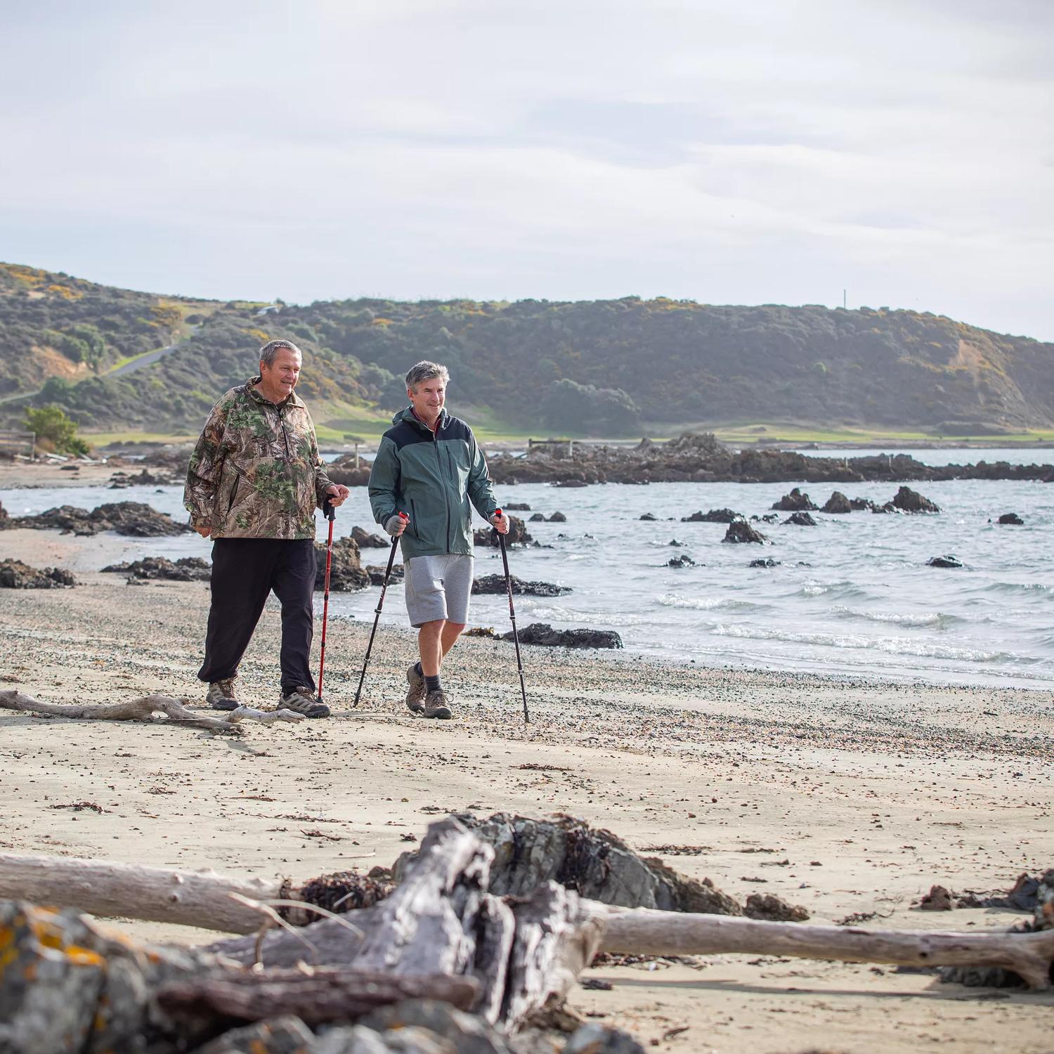Two people are using walking sticks to stroll along a beach.