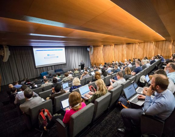 People sit in the auditorium at the Tiakiwai Conference Centre, watching the keynote speaker standing at the front