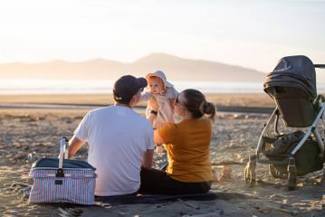 Two parents hold a baby in the air while having a picnic on a beach in the Kāpiti Coast, overlooking Kapiti Island.