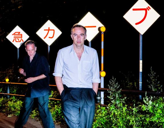 The two members of musical group 2manydjs, David and Stephen Dewaele, are on the side of the road with Japanese roadsigns at night.