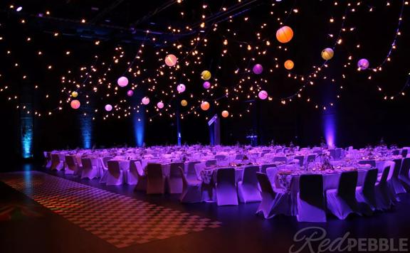 The interior of the St James Theatre located on Courtney Place in Wellington. The room is set up for a banquet with tables, with ten chairs at each and pink, purple and orange spherical lanterns suspended above.