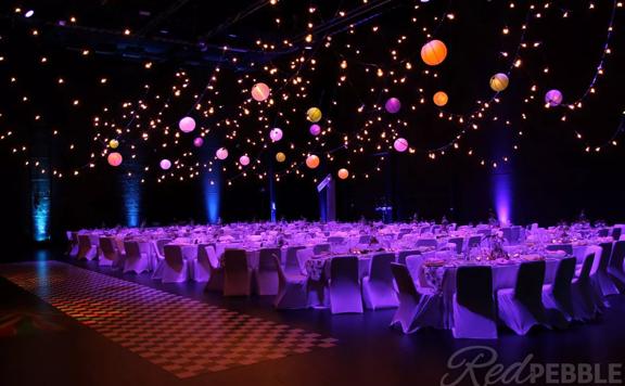 The interior of the St James Theatre located on Courtney Place in Wellington. The room is set up for a banquet with tables, with ten chairs at each and pink, purple and orange spherical lanterns suspended above. 