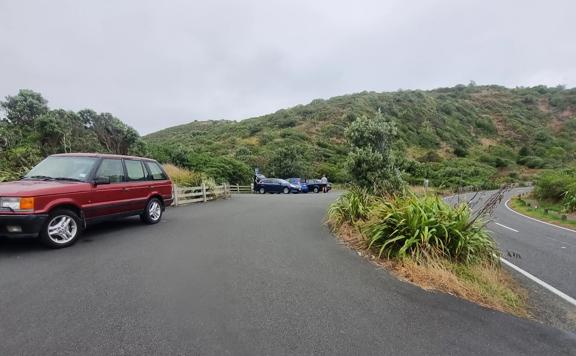 Cars parked in a flat car park with the main road on the right. The car park is surrounded by hills and native green bush.