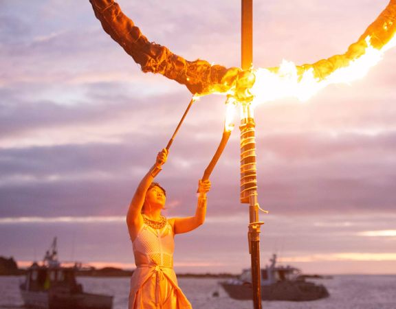 A person uses two torches to light a circular sculpture on fire for the Lōemis Festival. There is a body of water in the background with two parked boats and a pink-purply cloudy evening sky above.