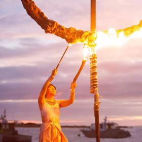 A person uses two torches to light a circular sculpture on fire for the Lōemis Festival. There is a body of water in the background with two parked boats and a pink-purply cloudy evening sky above. 