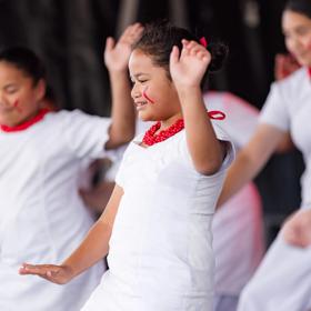 A group of Pasifika dancers perform on stage in white dresses and red beaded necklaces.