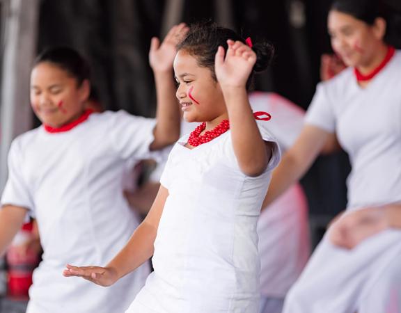 A group of Pasifika dancers perform on stage in white dresses and red beaded necklaces.