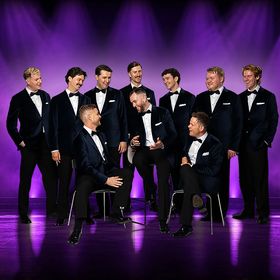 The Ten Tenors perform on stage under purple lighting, with ten singers in formal black suits arranged around seated and standing positions.