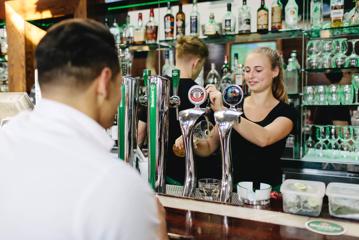 The bar at St Johns Bar & Eatery, a restaurant on Cable Street on Wellington's waterfront. A person wearing a white collared shirt is ordering a drink, two bartenders are behind the bar, one of them is pouring a draft beer into a large pint glass.