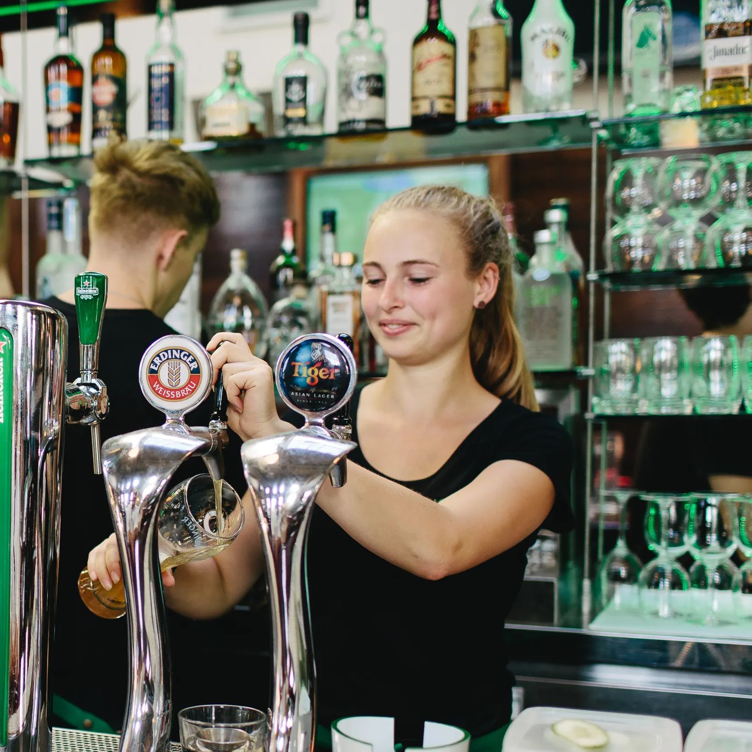 The bar at St Johns Bar & Eatery, a restaurant on Cable Street on Wellington's waterfront. A person wearing a white collared shirt is ordering a drink, two bartenders are behind the bar, one of them is pouring a draft beer into a large pint glass.