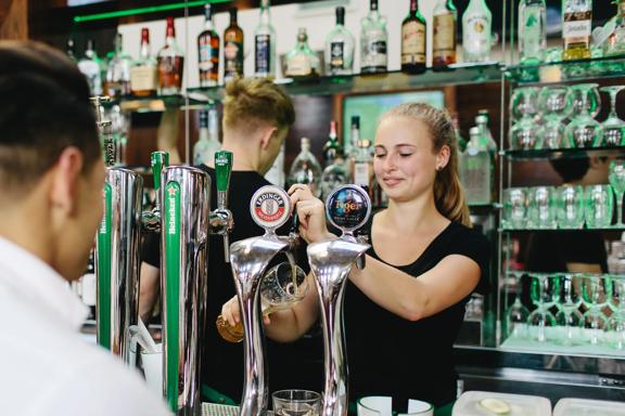 The bar at St Johns Bar & Eatery, a restaurant on Cable Street on Wellington's waterfront. A person wearing a white collared shirt is ordering a drink, two bartenders are behind the bar, one of them is pouring a draft beer into a large pint glass.