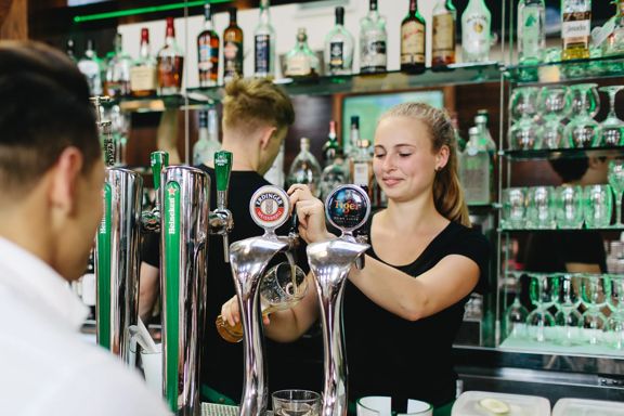 The bar at St Johns Bar & Eatery, a restaurant on Cable Street on Wellington's waterfront. A person wearing a white collared shirt is ordering a drink, two bartenders are behind the bar, one of them is pouring a draft beer into a large pint glass.