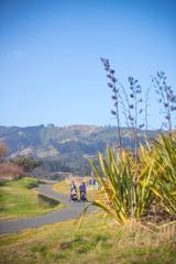 Two people, each pushing a push chair with a small child in it, walk along the sealed path in Queen Elizabeth park, Te ara o Whareroa.