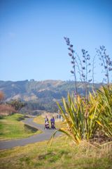 Two people, each pushing a push chair with a small child in it, walk along the sealed path in Queen Elizabeth park, Te ara o Whareroa.