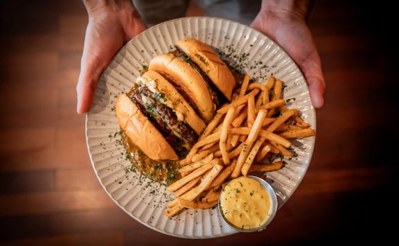 A plate held in two hands shows a burger alongside fries and a small cup of dipping sauce.