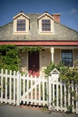 The front facade of Nairn Street Cottage in Wellington. The small off-white house has a red door, an over-grown yard and a white picket fence.