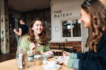 Two people sit at a table outside The Oatery in Left Bank. They are both eating from a bowl.