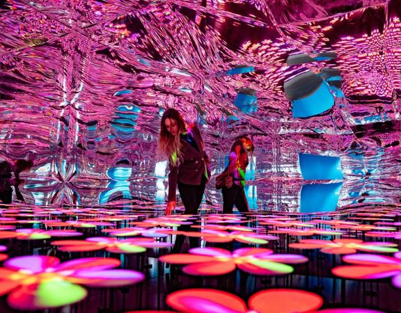 A person walks through an immersive art installation at the Whirinaki Whare Taonga, an arts centre in Upper Hutt Central. The space is illuminated in pink light from a flower-patterned floor and has a wavy, reflective ceiling.