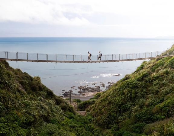 Two people walking along a swing bridge between two hillsides on the Escarpment track above the Kāpiti Coast.