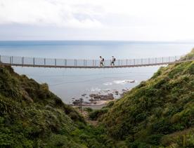 Two people walking along a swing bridge between two hillsides on the Escarpment track above the Kāpiti Coast.
