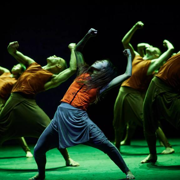 Akram Khan Company: Jungle Book reimagined, dancers are standing in a staggered formation with a wide stance, arms raised looking upward, illuminated by green-coloured light. 