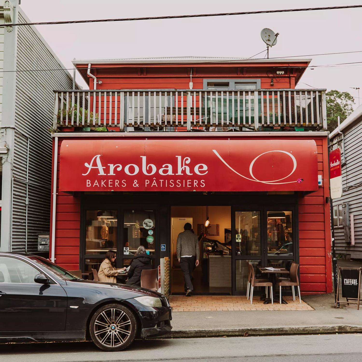 The front facade of Arobake Bakery in Aro Valley in Wellington. It is a small red-coloured two-story house with two four-seater tables outside.